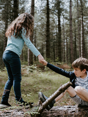 In a wooded area, a young girl helps a boy get up from a log. Image by Annie Spratt.