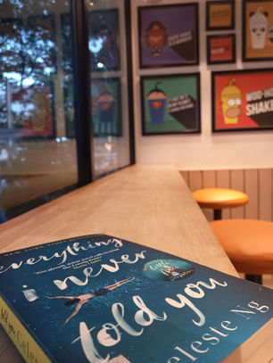 A copy of Everything I never told you by Celeste Ng lying on a wooden table and bar stools adjacent to a glass window. Photo by Ninay Desai.