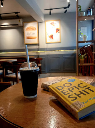 In a cafe, a copy of Do Epic Shit by Ankur Warikoo lies on a round, wooden table next to a frappe in a disposable cup. Photo by Ninay Desai.