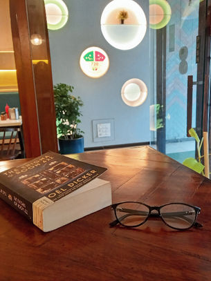 A copy of The Enigma of Room 622 by Joel Dicker and a pair of glasses lie on a wooden table in a cafe. Photo by Ninay Desai