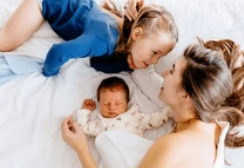 Woman and two children lie on a white bed. The woman and older child gaze lovingly at each other while the baby sleeps peacefully. Cozy mood.