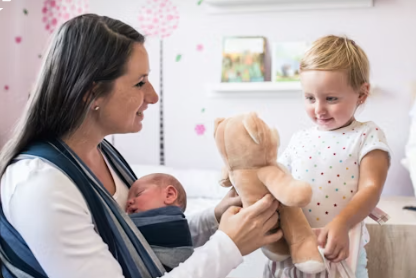 A woman holds a sleeping baby in a sling, smiling at a toddler with a teddy bear. Pink and white room with polka dot decor. Cozy and cheerful mood.