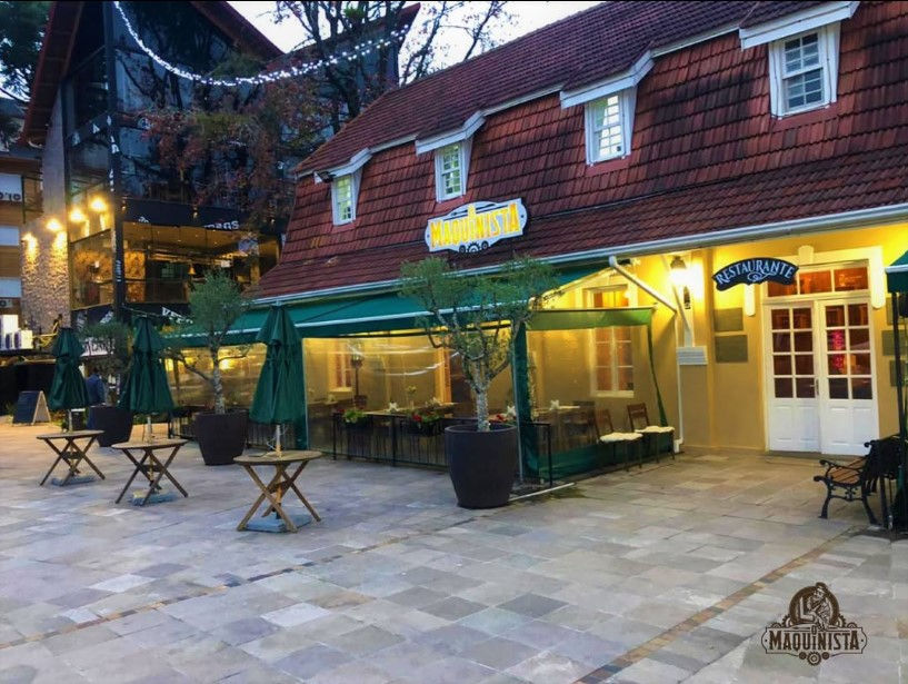 Fachada de restaurante com telhado vermelho e toldo verde. Mesas externas vazias e árvores em vasos. Ambiente tranquilo ao entardecer.