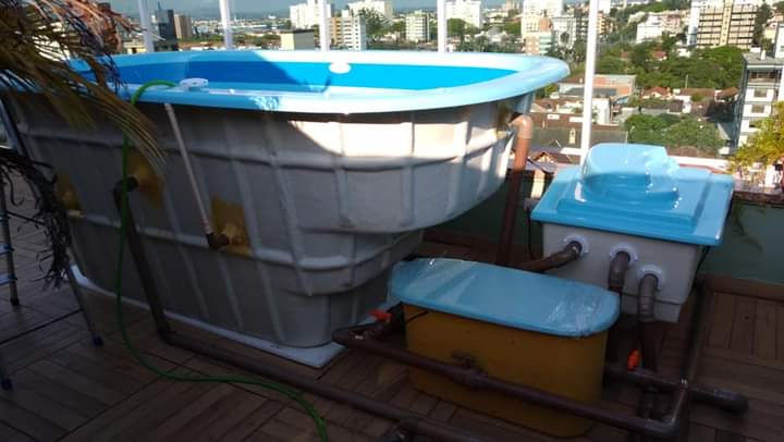 Piscina azul em varanda de cidade, cercada por sistemas de encanamento e caixas d'água. Ao fundo, prédios e céu claro.