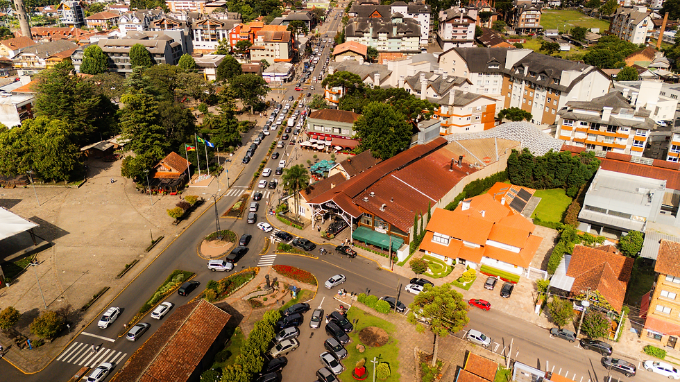 Vista aérea de uma cidade com muitos carros nas ruas, prédios ao fundo e uma praça arborizada. Telhados vermelhos se destacam. Dia ensolarado.