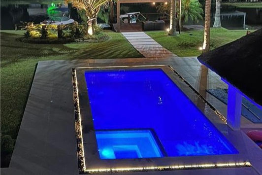 Piscina iluminada em azul à noite, cercada por palmeiras e grama. Ao fundo, uma área de estar à beira de um lago, com iluminação suave.