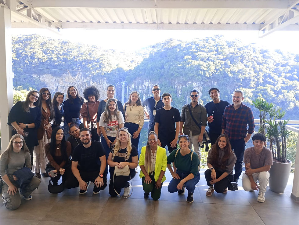 Grupo de 20 pessoas sorrindo em varanda com vegetação e cachoeira ao fundo. Ambiente claro, clima de alegria e amizade.