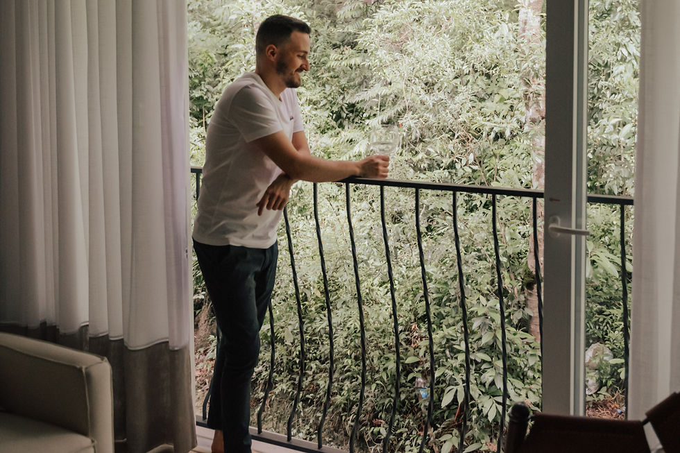 Homem de branco apoia-se na sacada, segurando um copo, olhando para a vista verde. Cortinas brancas ao lado, clima sereno e contemplativo.