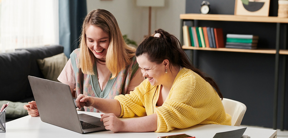 Two women sitting side by side, and smiling, as they look at the screen of an open laptop on the table in front of them.