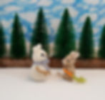 a photo of a small knitted mouse next to a snowman mouse with stick arms. The background is evergreen trees and cloudy blue skies.