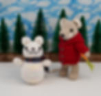 a photo of a knitted mouse in a red hoodie next to a snowman mouse with a scarf. In the background are evergreen trees and a cloudy blue sky.