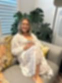 A smiling lady with a white flowy dress, sitting on a grey couch with a cup of tea. Ferns are behind her.