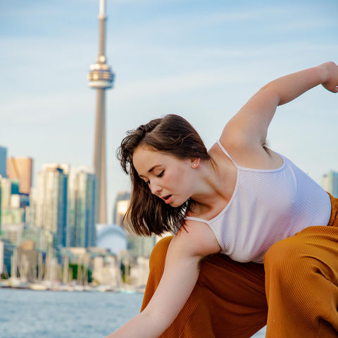 foreground: Dancer on a rock in crouched down, torso open tilt twist with arms open, head side profile looking down on an angle.
background dense city building with the CN tower and the sky.