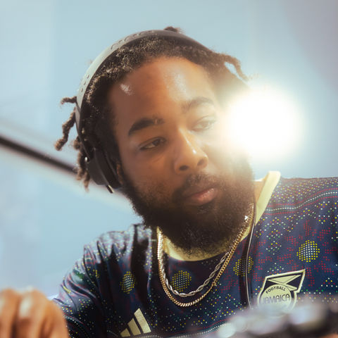 Headshot: head on a slight angle tilted to the side, gaze to the bottom corner. Hair in dreads, beard and mustache, relaxed face no smile. Wearing a pair of over-the-ear headphones, a few gold chains and a patterned shirt.