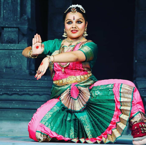Dancer posing kneeling on one knee with arms stretched out in front of the torso with one hand showing palm andone hand showing the back. Dressed in colourful pink, green and gold Traditional Indian outfit, smiling.