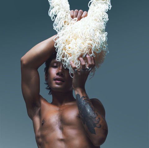 Portrait from hips up. Bare chest, crochet bottoms. Arms raised holding some pale yarn that partially covers an eye.