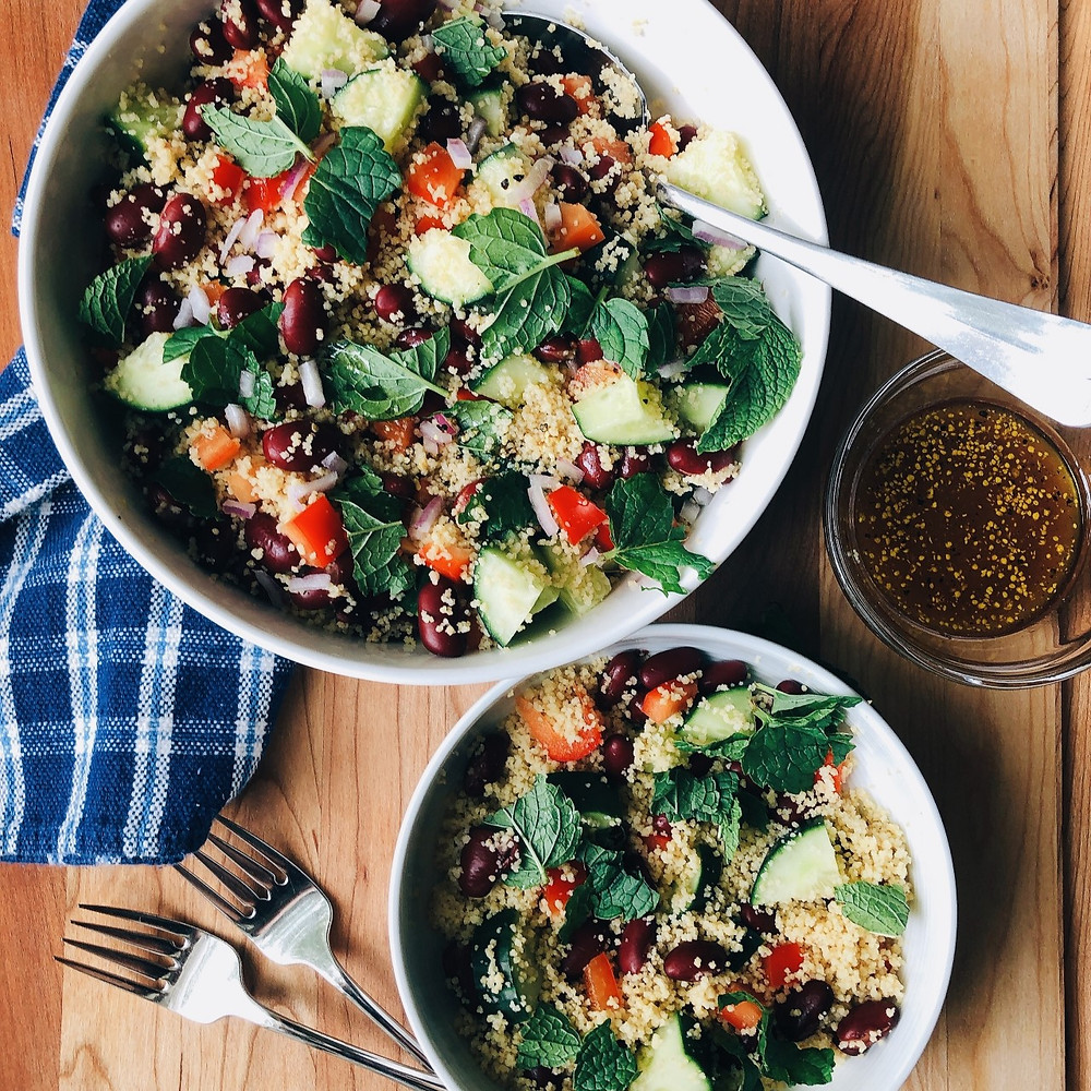 Three bowls sit on a wooden cutting board. Two bowls - one large salad bowl and one small serving bowl - are filled with whole wheat couscous with red wine vinegar dressing. The third bowl contains the rest of the red wine vinegar dressing. On the bottom left corner of the photo are two salad forks. The large salad bowl is wrapped with a blue and white paid tea towel.
