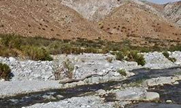 Alt text: Clear stream flowing over rocks through a rocky, arid desert landscape with dry hills and sparse vegetation under a partly cloudy sky.
