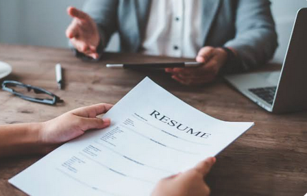 Image of a person's hand's holding a resume in front of another person.