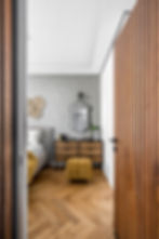 Modern bedroom with a wood door, patterned gray wall, and a decorative mirror. Features a yellow ottoman, cozy bed, and natural wood floor.