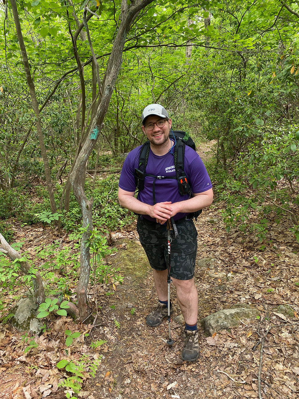 A white man stands next to teal hiking blaze in full hiking gear.