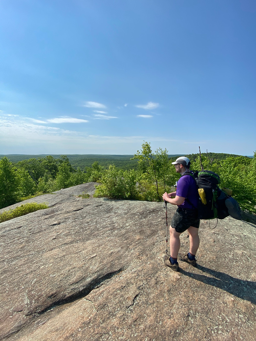 A white man in hiking gear stands on a rocky expanse and looks out at a vista on a sunny day