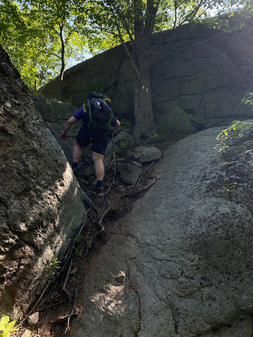 A white man hikes up a steep rocky gap between two larger boulders on a sunny day.