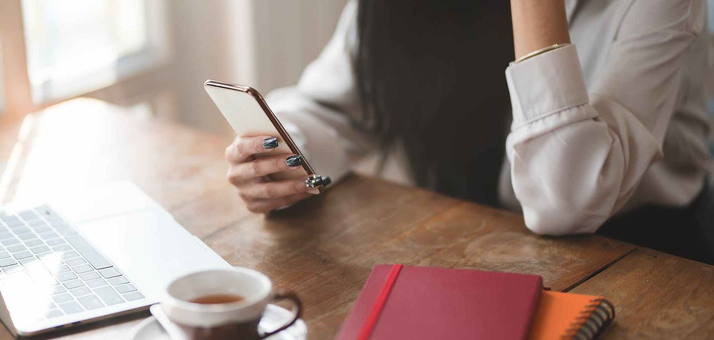 person holding black smartphone near white ceramic mug on brown wooden table