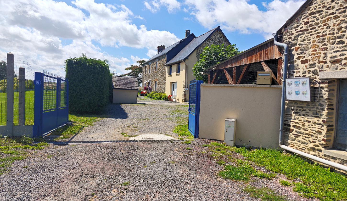 Photo de la propriété prise depuis la route. Sur la gauche de la photo, avant le portail bleu et au bord de la route, deux places de parking privées sont visibles. À l'intérieur de la propriété, sur la gauche, se trouve le jardin bordé de thuyas, séparant la cour d'une partie du jardin. Sur la droite de la photo, une partie d'un bâtiment secondaire (ancien poulailler et écurie) situé au niveau de la route. Ce bâtiment possède des panneaux "Chambres d'Hôtes Les Salles" et "parking" de chaque côté, ainsi qu'un tableau d'affichage mural présentant les trois chambres avec leurs tarifs et un QR code menant à une page de réservation et de vérification des disponibilités. Au centre de la photo, un portail bleu, derrière lequel se trouve la cour avec une place de stationnement devant une petite maisonnette. En arrière-plan, le bâtiment principal est visible.