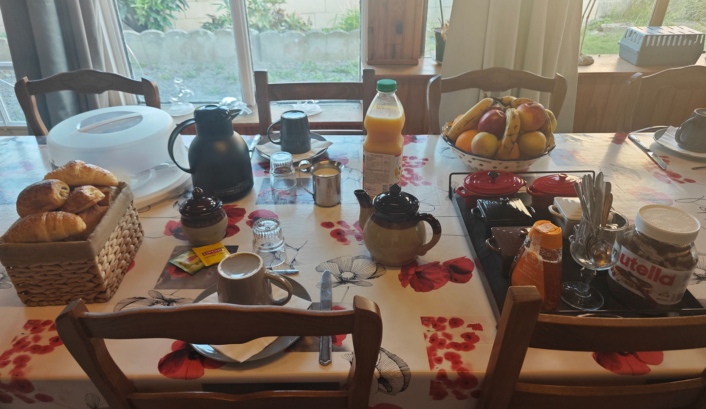 Le petit-déjeuner est présenté sur une grande table dans la véranda. Il n'est pas servi sous forme de buffet ; tous les éléments sont disposés sur la table pour que les clients se servent.
Placement et Vaisselle : Les tables sont dressées avec des assiettes, des couverts (couteau, cuillère à café), des mugs et des verres.
Boissons chaudes et froides : Vous trouverez sur la table une carafe de café, une théière d'eau chaude, du thé (vert et noir), un petit pichet de lait et du jus d'orange. Du chocolat chaud en poudre et du lait chaud sont disponibles sur demande.
Pains et viennoiseries : Une bannette contient du pain frais, des mini-croissants et des mini-pains au chocolat.
Produits laitiers et tartinables : Sous une cloche se trouvent des fromages (Saint-Môret, Babybel, La Vache qui Rit), du beurre doux et des yaourts. Vous trouverez également du miel, du Nutella et un assortiment de confitures maison (servies dans des petites cocottes) ainsi que des confitures classiques.
Fruits : Une corbeille de fruits frais est à votre disposition avec des pommes, des oranges et des bananes.