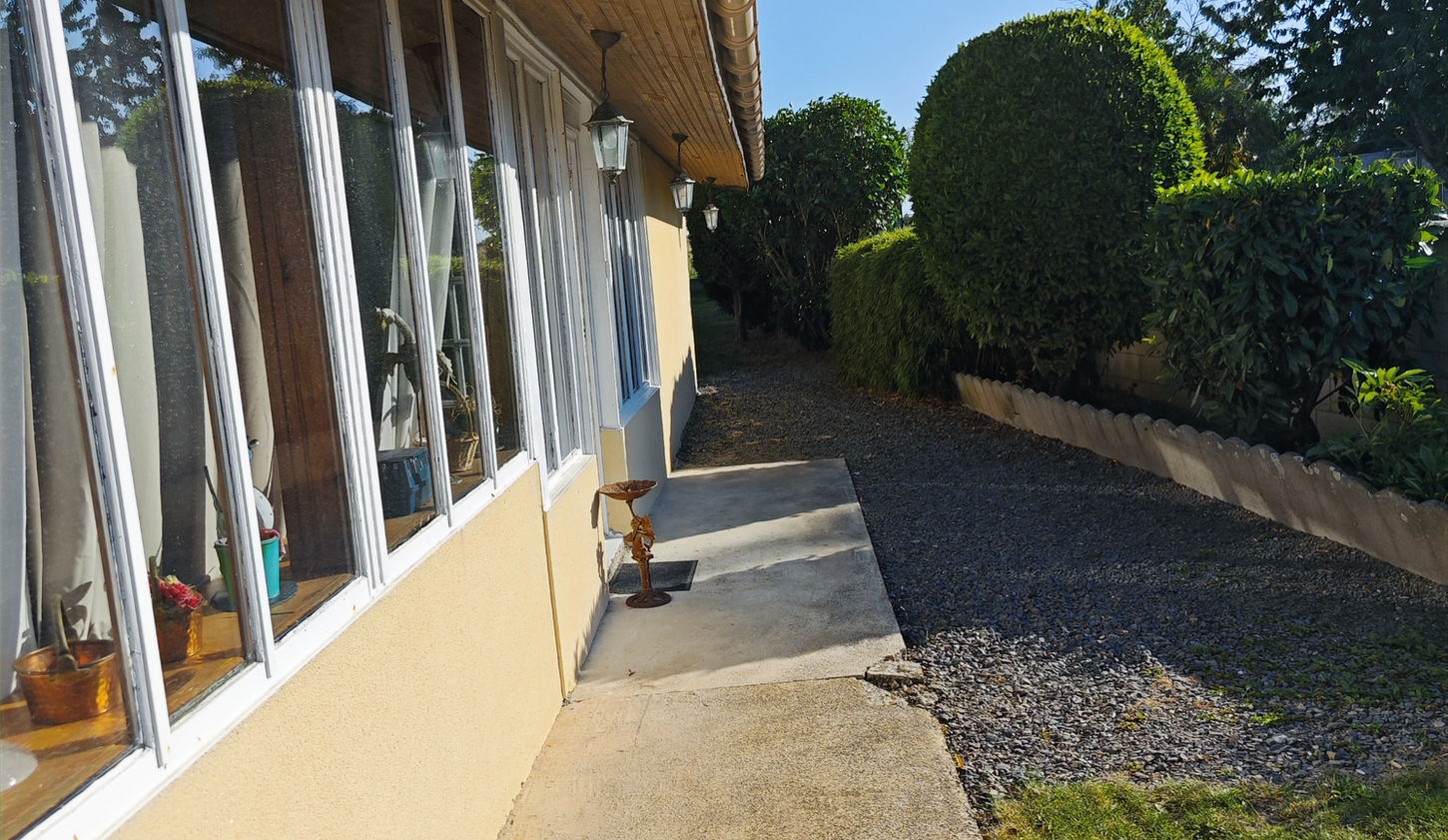 Photo de la véranda des Chambres d'Hôtes "Les Salles", prise depuis le chemin qui y mène. À gauche, les fenêtres et la longue façade de la véranda sont visibles ; elle est située à l'arrière du bâtiment principal et sert de réception et de salle de petit-déjeuner. À droite, on aperçoit une partie de la cour et une partie du jardin avec des plates-bandes d'arbustes, derrière lesquelles se trouve un mur mitoyen avec la propriété voisine.