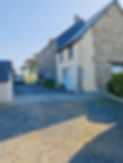 Photo extérieure du bâtiment principal des Chambres d'Hôtes "Les Salles", prise depuis l'entrée de la propriété. Au premier plan, la cour avec des places de parking. Sur la gauche, une petite maisonnette (ancienne maison du chien) avec une place de parking devant. À droite, des arbustes et un petit panneau "Réception" indiquant un chemin longeant le bâtiment principal. La façade visible présente du crépi jaune, deux fenêtres à l'étage avec ferronnerie bleue (pour la Chambre à Fleurs, ancienne pigeonnier), et en dessous, deux portes indépendantes (Chambre Capucine et Lilas, ancienne salle de jeu/garage). Une autre porte d'entrée sur cette façade peut être utilisée par les clients. La porte d'entrée principale pour les clients de la Chambre à Fleurs est celle de la véranda, située de l'autre côté du bâtiment principal. Exterior photo of the main building of "Les Salles" Bed & Breakfast, taken from the property's entrance. In the foreground, the courtyard with parking spaces. On the left, a small doghouse with a parking spot in front. To the right, shrubs and a small "Reception" sign indicating a path alongside the main building. The visible facade features yellow rendering, two upstairs windows with blue wrought ironwork (for the Flower Room, former dovecote), and below, two independent doors (Capuchin Room and Lilac Room, former playroom/garage). Another entrance door on this facade can be used by guests. The main entrance for Flower Room guests is via the veranda, located on the other side of the main building.