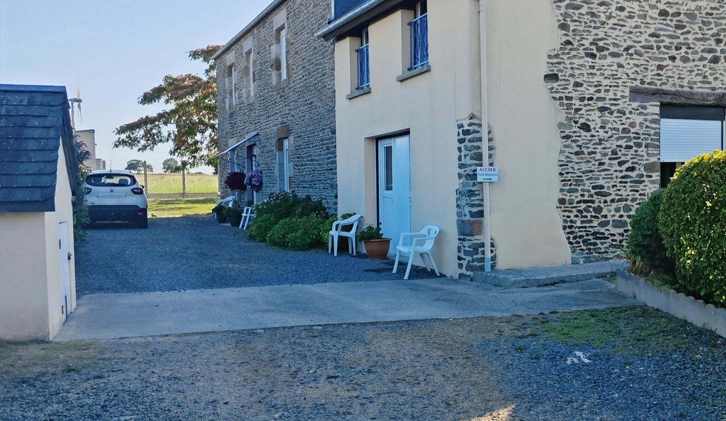 Photo extérieure du bâtiment principal des Chambres d'Hôtes "Les Salles", prise depuis l'entrée de la propriété. Au premier plan, la cour avec des places de parking. Sur la gauche, une petite maisonnette (ancienne maison du chien) avec une place de parking devant. À droite, des arbustes et un petit panneau "Réception" indiquant un chemin longeant le bâtiment principal. La façade visible présente du crépi jaune, deux fenêtres à l'étage avec ferronnerie bleue (pour la Chambre à Fleurs, ancienne pigeonnier), et en dessous, deux portes indépendantes (Chambre Capucine et Lilas, ancienne salle de jeu/garage). Une autre porte d'entrée sur cette façade peut être utilisée par les clients. La porte d'entrée principale pour les clients de la Chambre à Fleurs est celle de la véranda, située de l'autre côté du bâtiment principal.