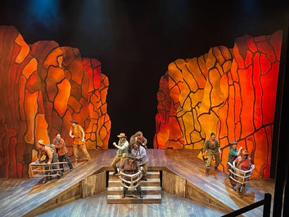 Red cliffs surround actors on two ramps and stairs, in outlines of boats