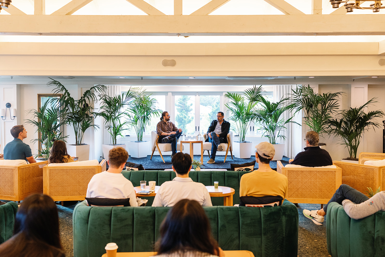 Audience on green sofas watching a two-person panel in a bright room with palms