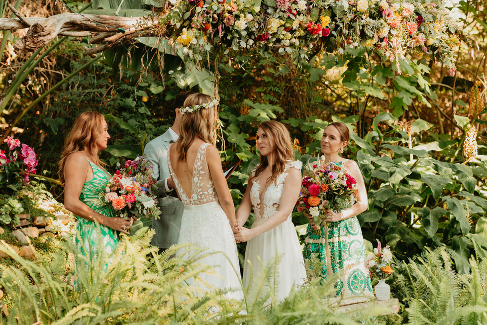 Brides hold hands during their wedding ceremony under an abundance of bright flowers