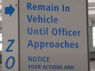 Sign at a U.S. Port of Entry instructing drivers to remain in their vehicles until an officer approaches. The sign also notifies that actions and conversations are recorded.