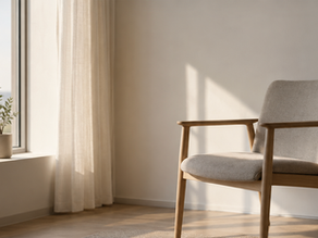 A very minimal photograph of an empty chair beside a window, soft natural light, lots of negative space, muted neutral tones.