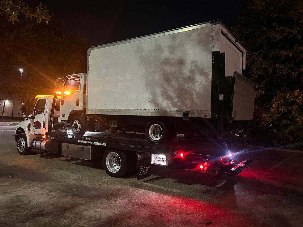 Medium Duty Tow truck hauling a white box truck at night, amber lights flashing on a dark street, with trees in the background.