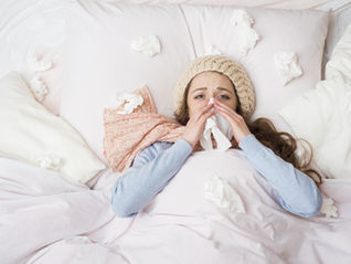 A young woman lies sick in bed with a wool hat and scarf, blowing her nose into a tissue. Crumpled tissues are scattered around her on the bed, indicating she has a cold or flu.