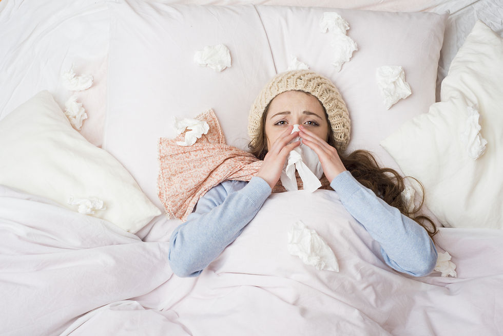A young woman lies sick in bed with a wool hat and scarf, blowing her nose into a tissue. Crumpled tissues are scattered around her on the bed, indicating she has a cold or flu.