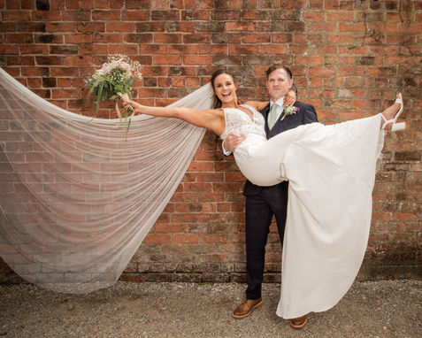 A colour photograph of a groom carrying his bride who is holding her bouquet out.