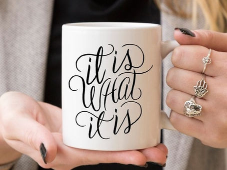 Hands hold a white mug with cursive text: "It is what it is." Person wears rings, nails painted black; background is neutral.
