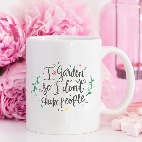 White mug with floral text reads "I garden so I don't choke people" in front of pink flowers. Pink accents create a humorous mood.