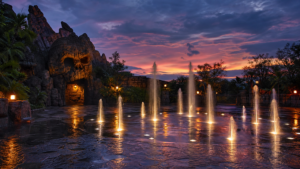 Illuminated fountains in a theme park plaza at sunset, facing a rocky formation carved with a skull-like face, with a castle in the background.