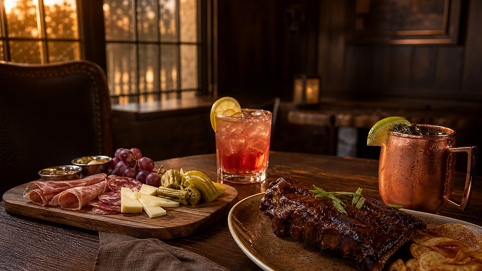 Una mesa de comedor tenue con una tabla de carnes frías, quesos, uvas y pepinillos, junto a un cóctel con rodaja de limón, una taza de cobre y un plato de costillas con guarnición.