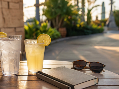 Mesa de madera al aire libre con dos bebidas frías con rodajas de limón, un libro cerrado y gafas de sol; al fondo se ve un camino soleado con vegetación y estructuras desenfocadas.