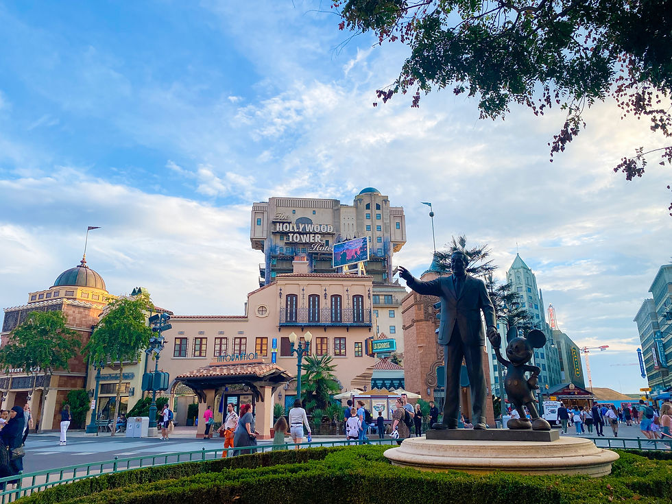 Statue of Walt Disney and Mickey Mouse in a plaza with people walking. Background features "Hollywood Tower Hotel" building, blue sky above.