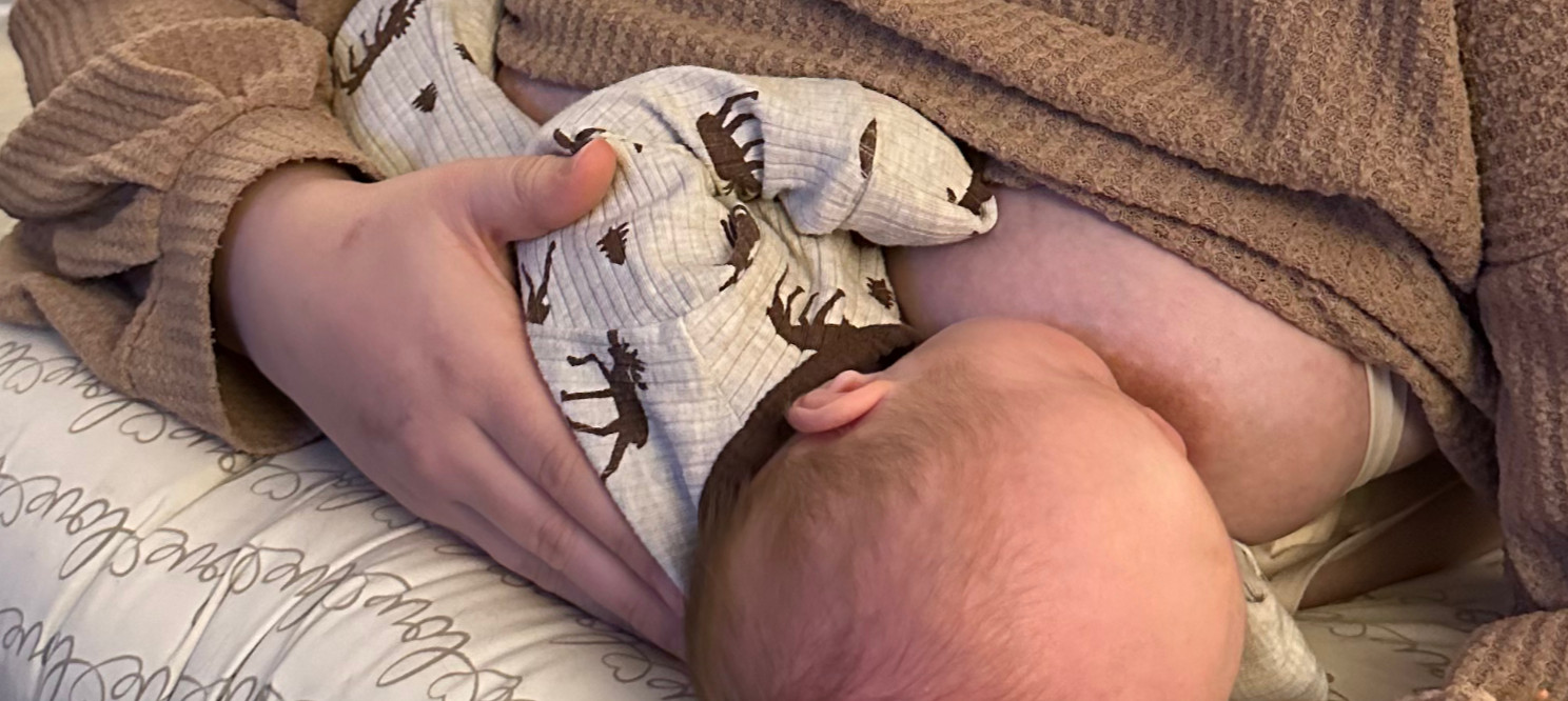 Baby nursing in a moose-print onesie cradled in an adult’s arms on a scripted pillow, cozy brown robe background