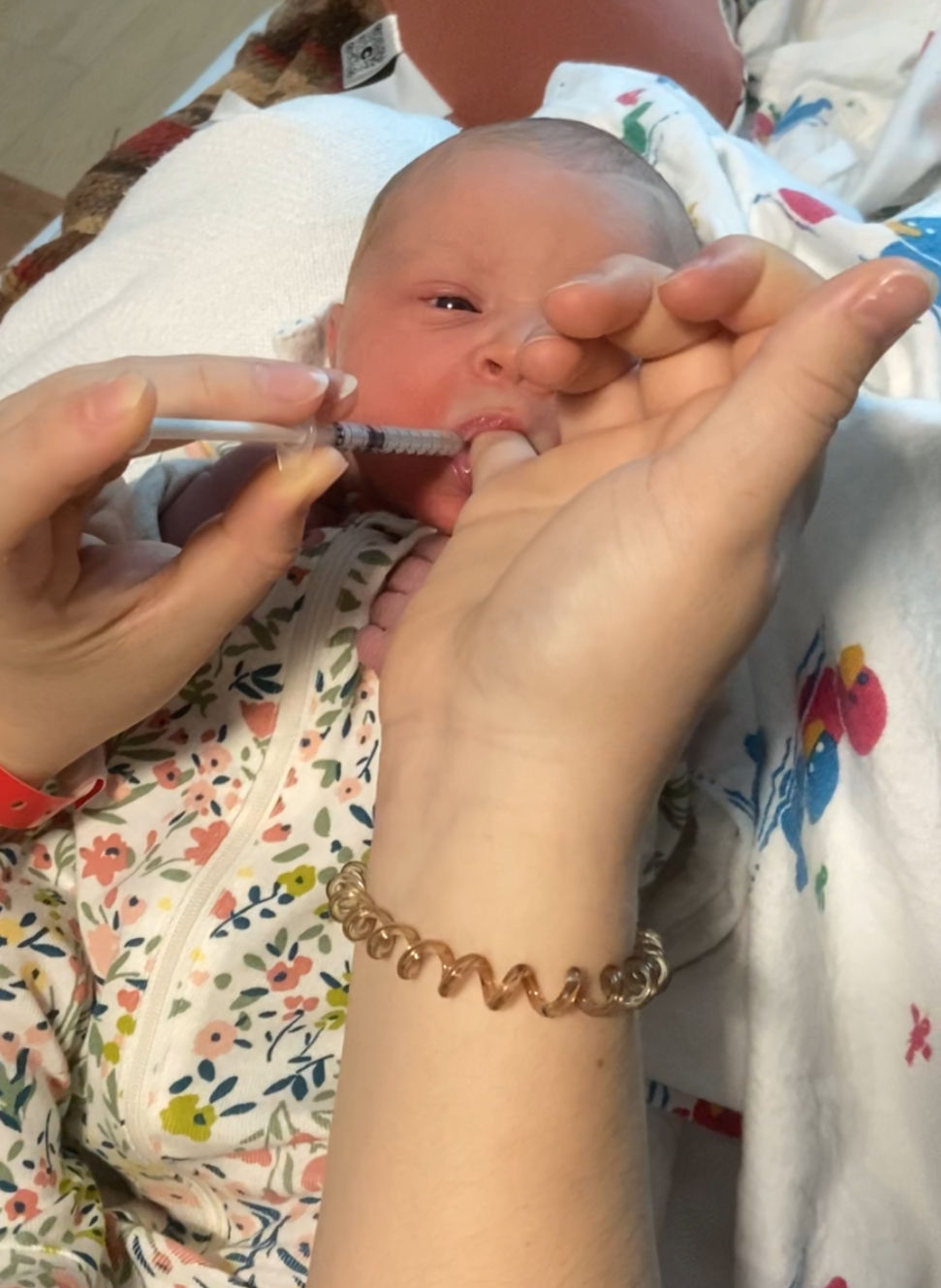 Adult hands feed a newborn colostrum with a syringe, baby in a floral onesie on a bed, looking sleepy.